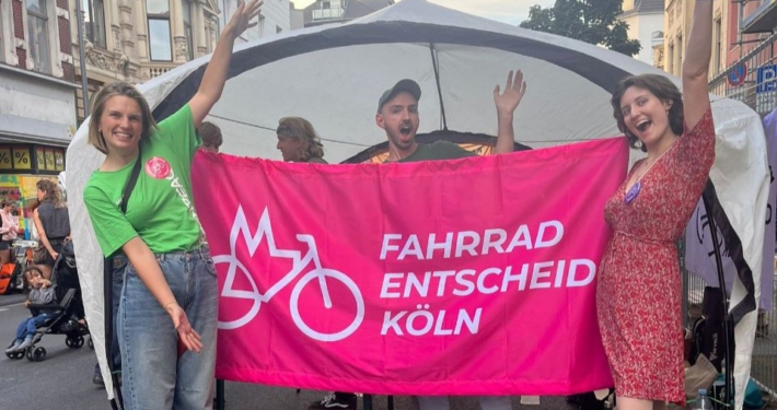 Drei Leute vom Fahrrad-Entscheid halten beim Bonner Straßenfest ein Banner mit unserem Logo hoch.