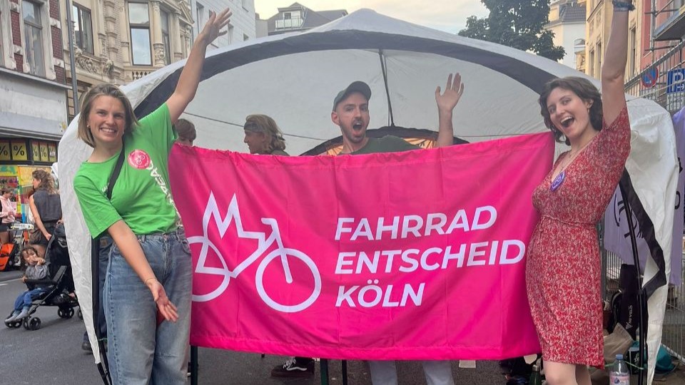 Drei Leute vom Fahrrad-Entscheid halten beim Bonner Straßenfest ein Banner mit unserem Logo hoch.